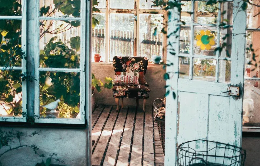 Cabane de jardin à l'ancienne, portes vitrées bleu pâle ouvertes sur un fauteuil fleuri
